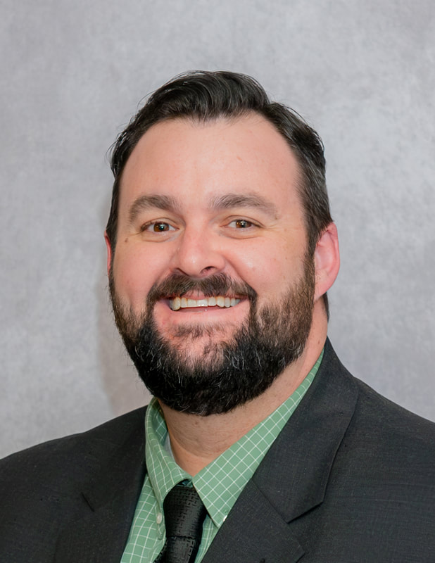 A man with a beard and mustache smiles at the camera, wearing a black suit and a green checkered shirt.