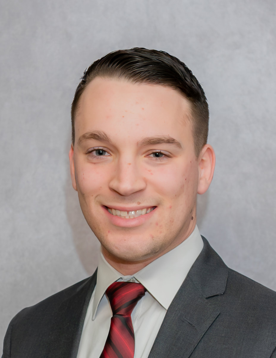 A smiling man in a suit and tie poses for a professional headshot against a gray background.