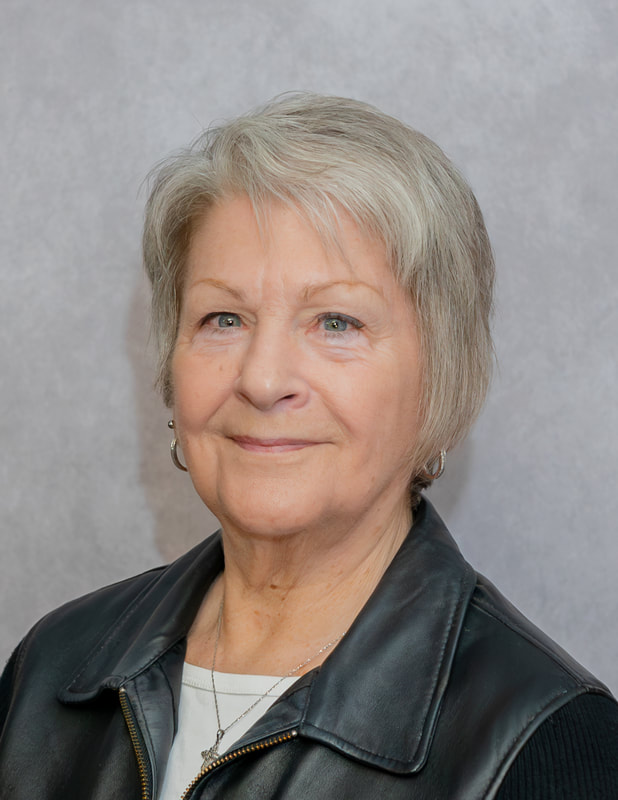 A senior woman with short, light-colored hair, wearing a black leather jacket, smiles softly against a neutral gray background.