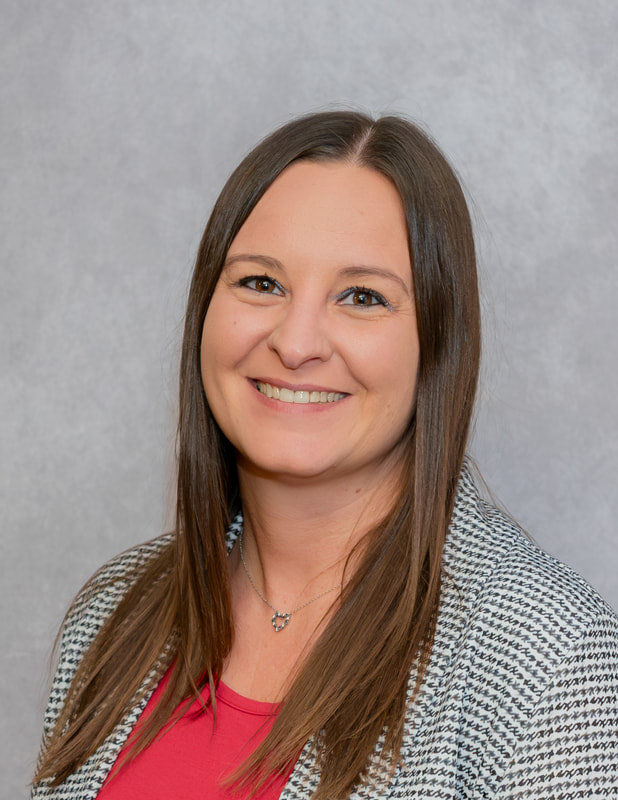 A woman with long brown hair, wearing a red top and a patterned blazer, smiles against a gray background.