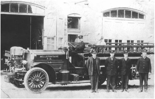 A historic black and white photograph featuring a vintage fire truck parked in front of a fire station, with four men in formal attire standing beside it, and one man seated in the driver's seat.