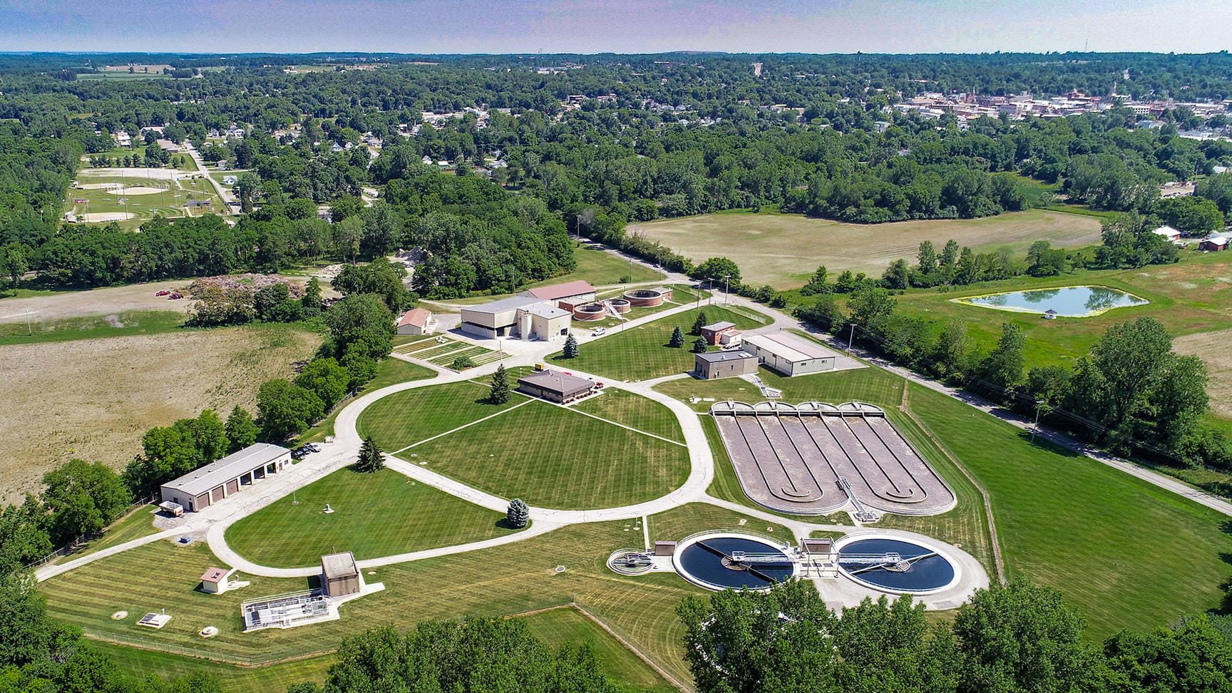 An aerial view of a water treatment facility surrounded by green fields and trees, featuring circular and rectangular tanks for water processing.