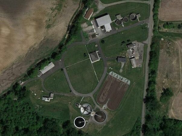 An aerial view of a facility featuring several buildings, circular structures, and open green spaces, surrounded by agricultural land.