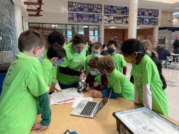 A group of children in green t-shirts gather around a table, intently observing a small robot while using a laptop in a school setting.