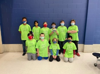 A group of ten children wearing bright green t-shirts and masks, posing together in front of a blue wall.