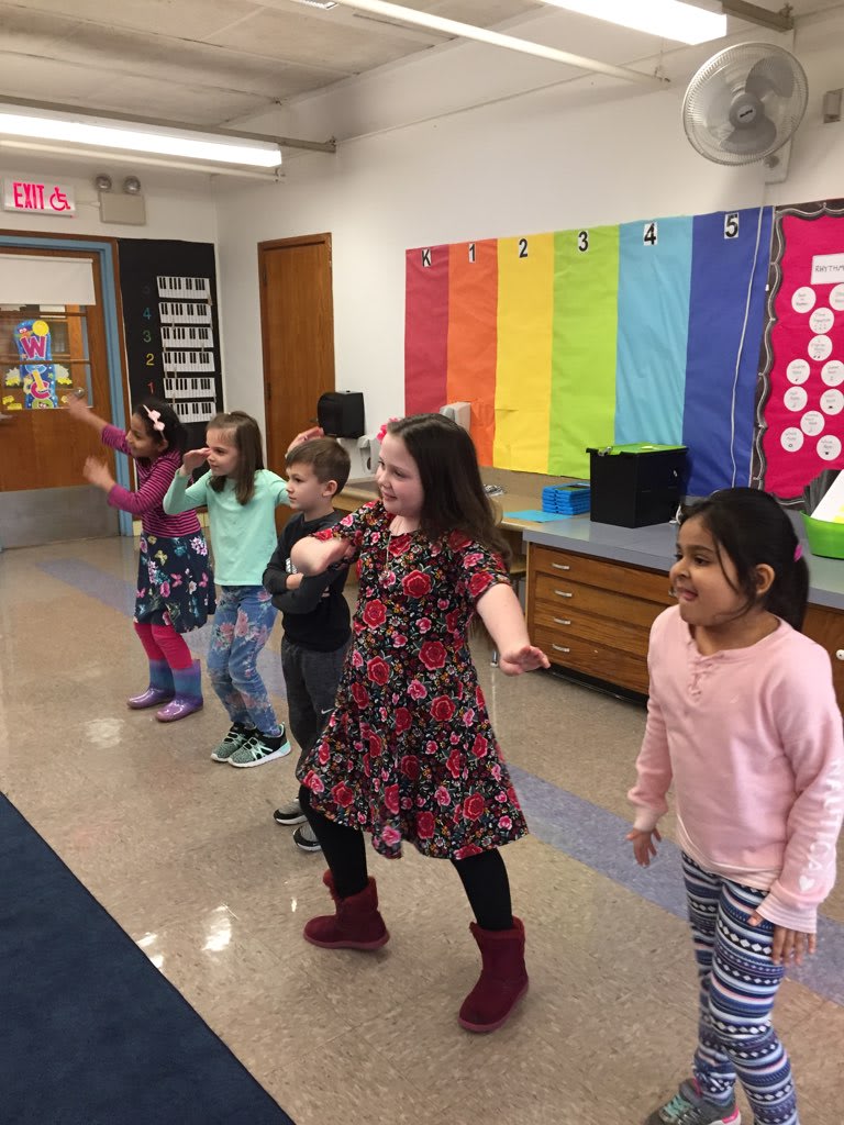 A group of five children, dressed in colorful outfits, are engaged in a dance activity in a classroom with bright colored walls and a carpeted floor.