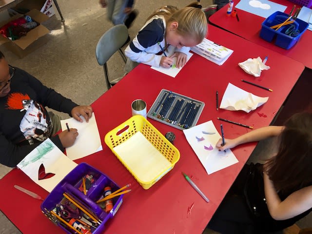 A top-down view of a classroom table where three children are engaged in art activities, using colored pencils and paper, with various art supplies organized in containers.