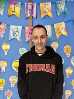 A person wearing a black hoodie with 'THOMAS' printed in red stands in front of a colorful classroom backdrop featuring banners that read 'FIRST GRADE' and various decorative hot air balloons.