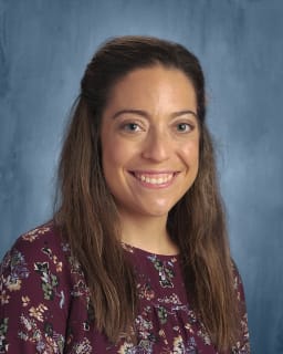 A smiling woman with long brown hair, wearing a floral blouse, poses against a blue background.