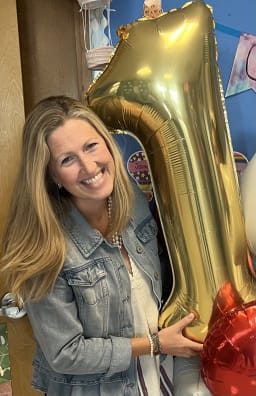 A smiling woman holds a large gold number '1' balloon, surrounded by colorful decorations in a cheerful setting.
