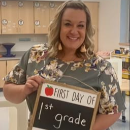 A smiling woman holds a chalkboard sign that reads 'FIRST DAY OF 1st grade' with a red apple graphic, standing in a classroom setting.