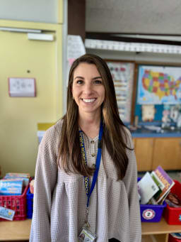 A smiling woman with long brown hair stands in a classroom, wearing a light-colored blouse and a lanyard with an ID badge, with bookshelves and a map visible in the background.