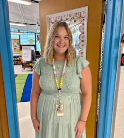 A smiling woman stands in a classroom doorway, wearing a light green dress and a colorful beaded necklace, with a classroom visible in the background.