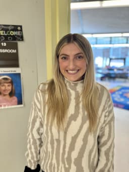 A smiling woman with long blonde hair wearing a striped sweater stands in a brightly lit hallway next to a classroom door labeled 'Welcome! Classroom 12'.