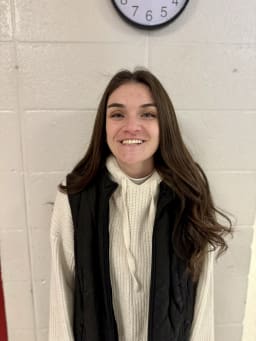A young woman with long brown hair is smiling while wearing a black vest over a white sweater, standing in front of a white wall with a clock above her.