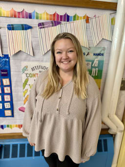A smiling woman with long hair stands in front of a colorful classroom bulletin board decorated with banners and educational posters.