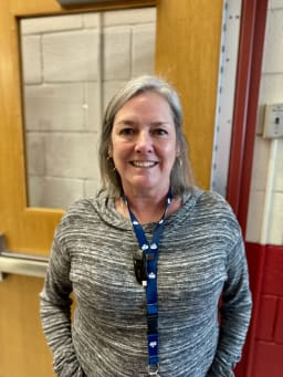 A smiling woman with shoulder-length gray hair, wearing a gray sweater and a blue lanyard, stands in front of a wooden door with a window.