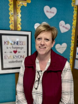 A smiling woman with short blonde hair stands in front of a colorful classroom wall decorated with hearts and motivational quotes about kindness and inclusion.