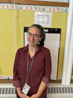 A smiling woman with glasses stands in front of a classroom wall decorated with colorful trim, wearing a maroon shirt and a lanyard with an ID badge.