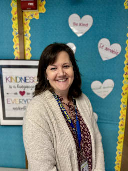 A smiling woman stands in front of a colorful classroom wall decorated with hearts and a poster promoting kindness.