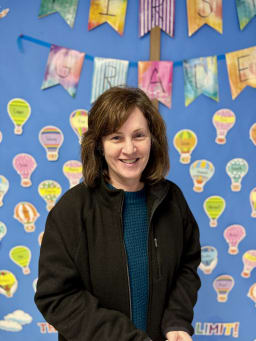 A smiling woman stands in front of a colorful classroom wall decorated with hot air balloon drawings and banners that read 'First Grade'.