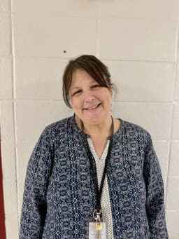 A woman with brown hair, wearing a patterned blue cardigan over a white top, smiles while standing against a white wall.
