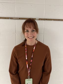A smiling woman with red hair, wearing a brown sweater and a lanyard, stands in front of a white wall.