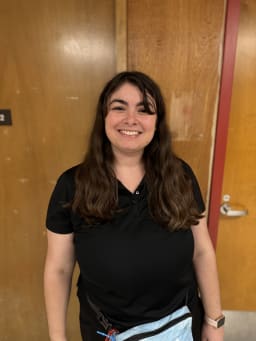 A smiling woman with long brown hair stands in front of a wooden wall, wearing a black polo shirt and a light blue bag across her waist.