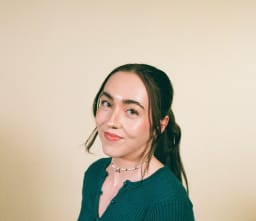 A young woman with long hair tied back, wearing a green top and a choker necklace, smiles softly against a plain beige background.
