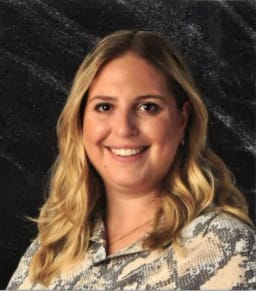 A smiling woman with long, wavy blonde hair, wearing a patterned shirt, poses against a dark textured background.