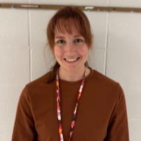 A smiling woman with red hair, wearing a brown sweater and a lanyard, stands in front of a white wall.