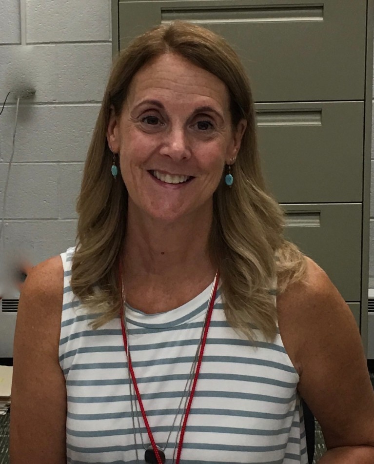 A smiling woman with long, wavy hair wearing a striped sleeveless top and earrings, sitting in front of a filing cabinet.