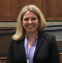 A woman with shoulder-length blonde hair, wearing a black blazer over a purple top, smiles while standing in a venue with wooden paneling in the background.