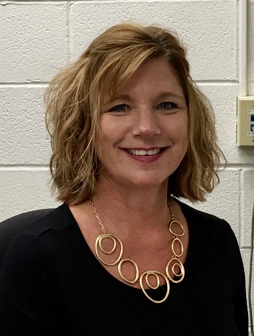A woman with shoulder-length wavy hair smiles while wearing a black top and a gold circular necklace, standing in front of a light-colored wall.