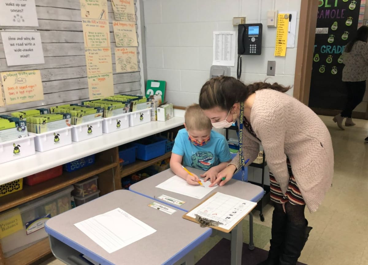 A teacher assists a young student with a writing task at a classroom table, surrounded by educational materials and supplies.