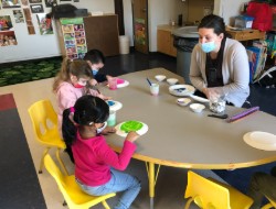 A teacher and four children, all wearing masks, are engaged in a painting activity at a round table in a classroom, with various paint supplies and plates in front of them.