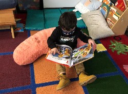 A young child sits on a colorful carpet, reading a magazine while resting against a large, soft pillow.
