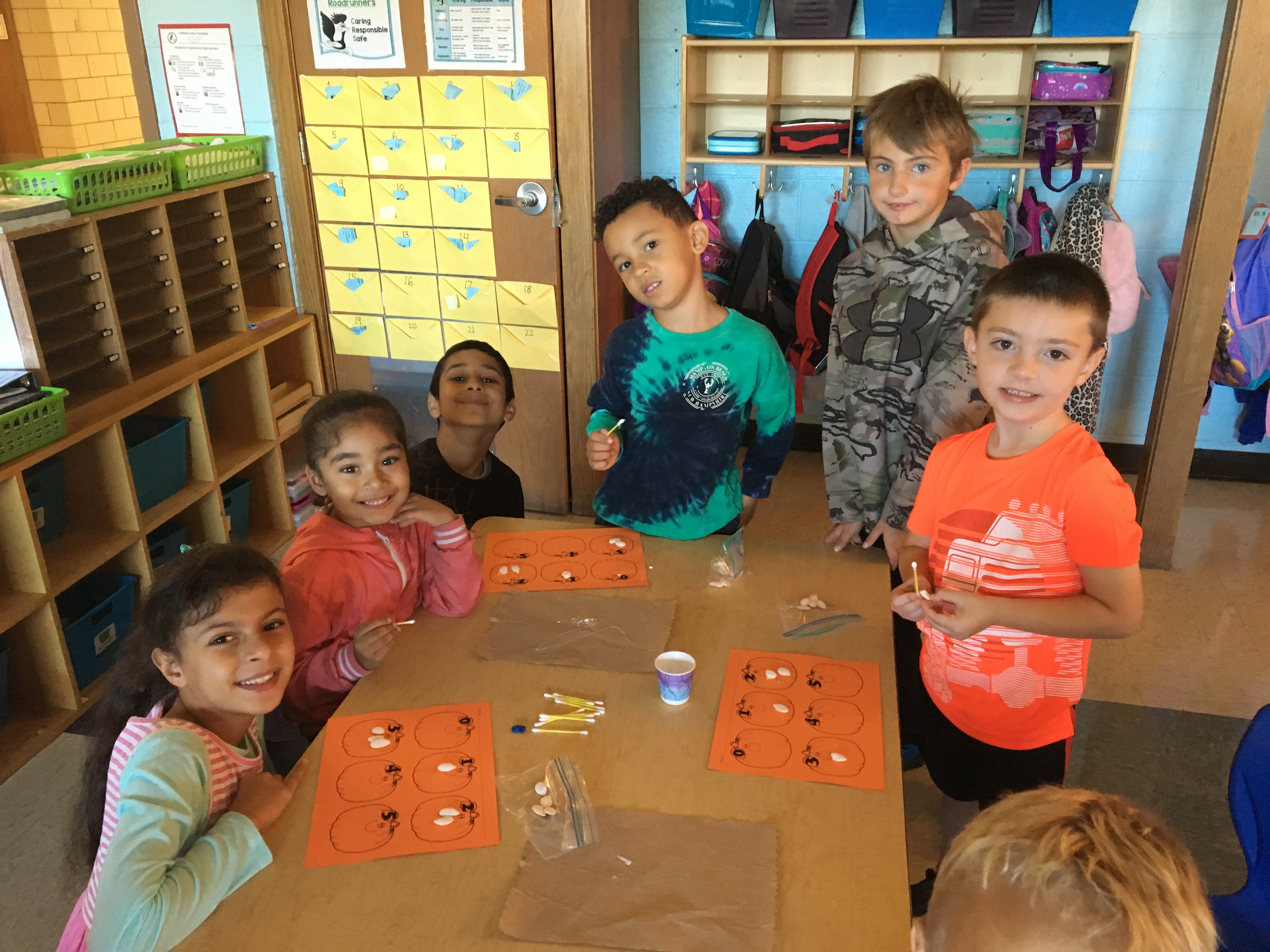 A group of six children, engaged in a classroom activity, are gathered around a table with orange sheets and various materials, smiling at the camera.