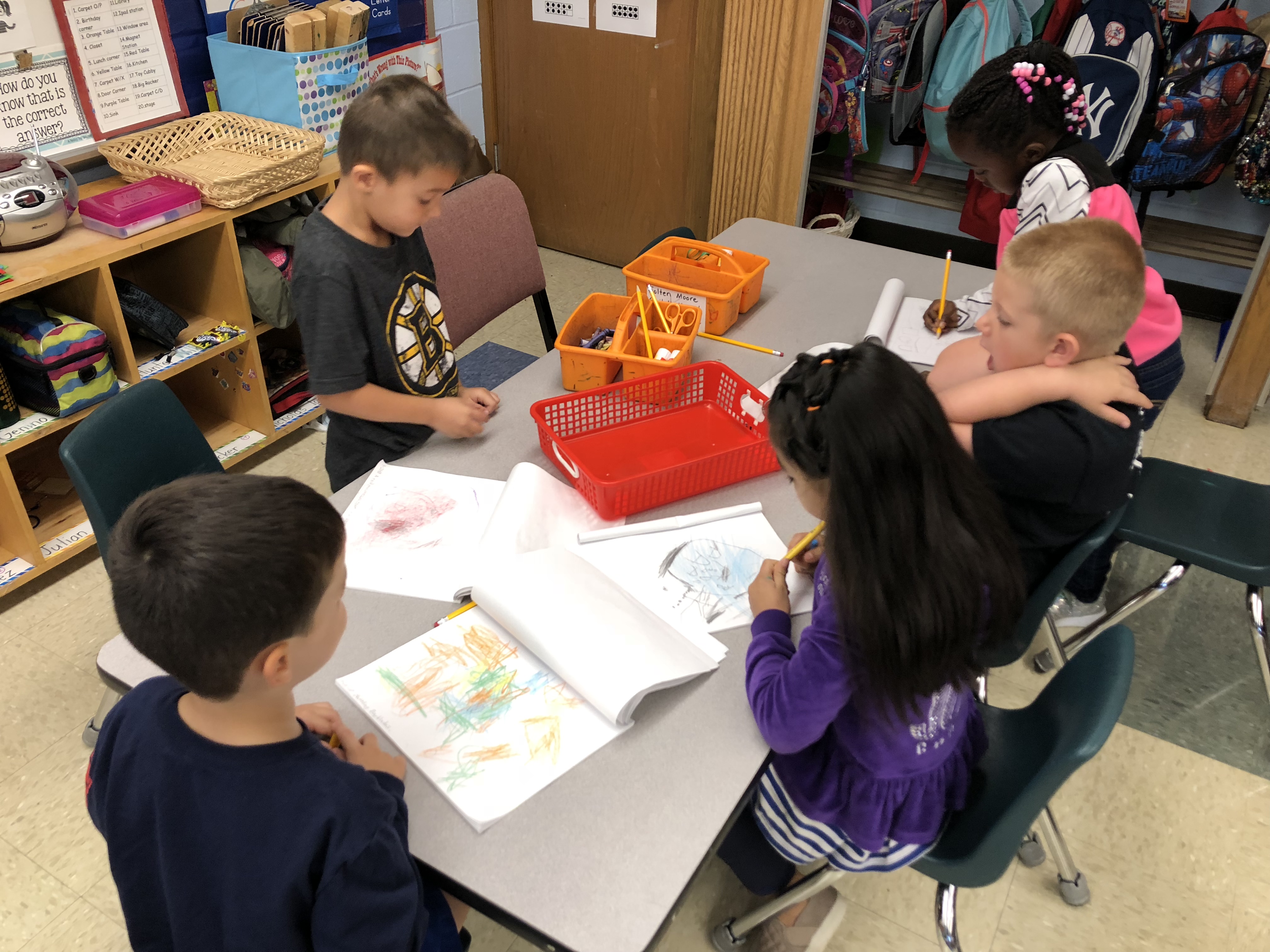 A group of five young children are seated around a table, engaged in coloring and drawing activities, with art supplies and their drawings spread out in front of them.