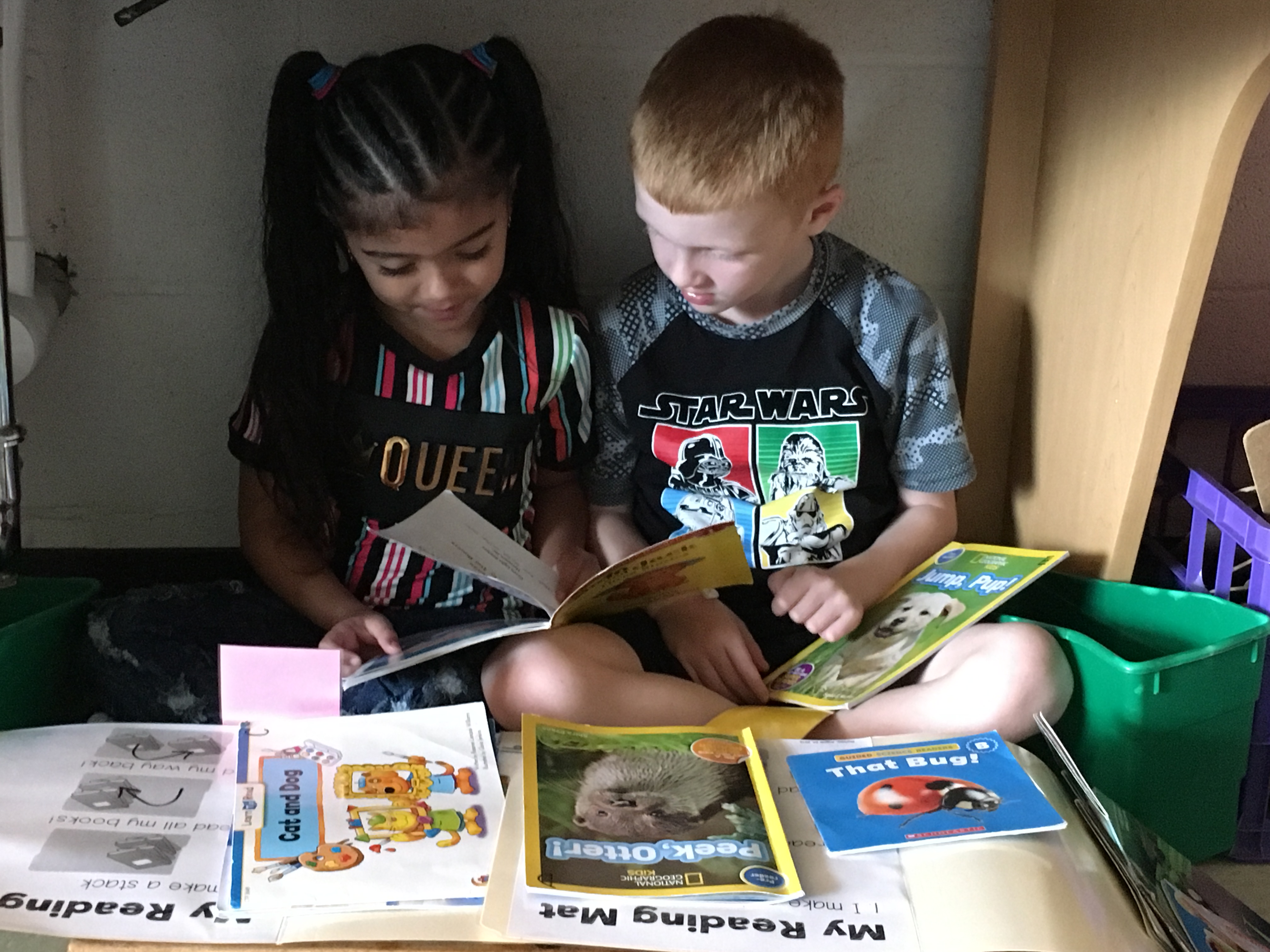 Two children, a girl with long braided hair and a boy with short red hair, sit together in a cozy reading nook surrounded by colorful children's books, as they share a story.