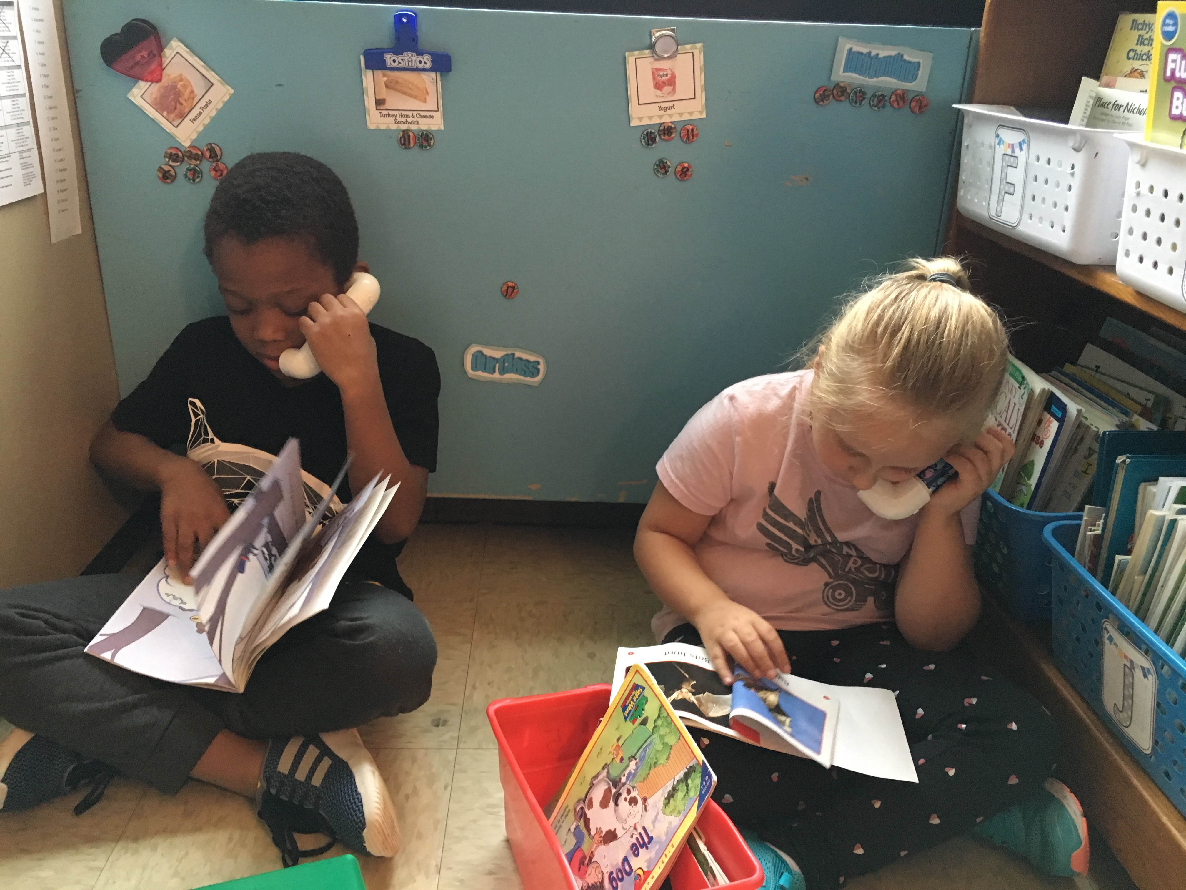 Two children are sitting on the floor in a cozy reading nook, each holding a book and a toy phone to their ears, engaged in imaginative play.