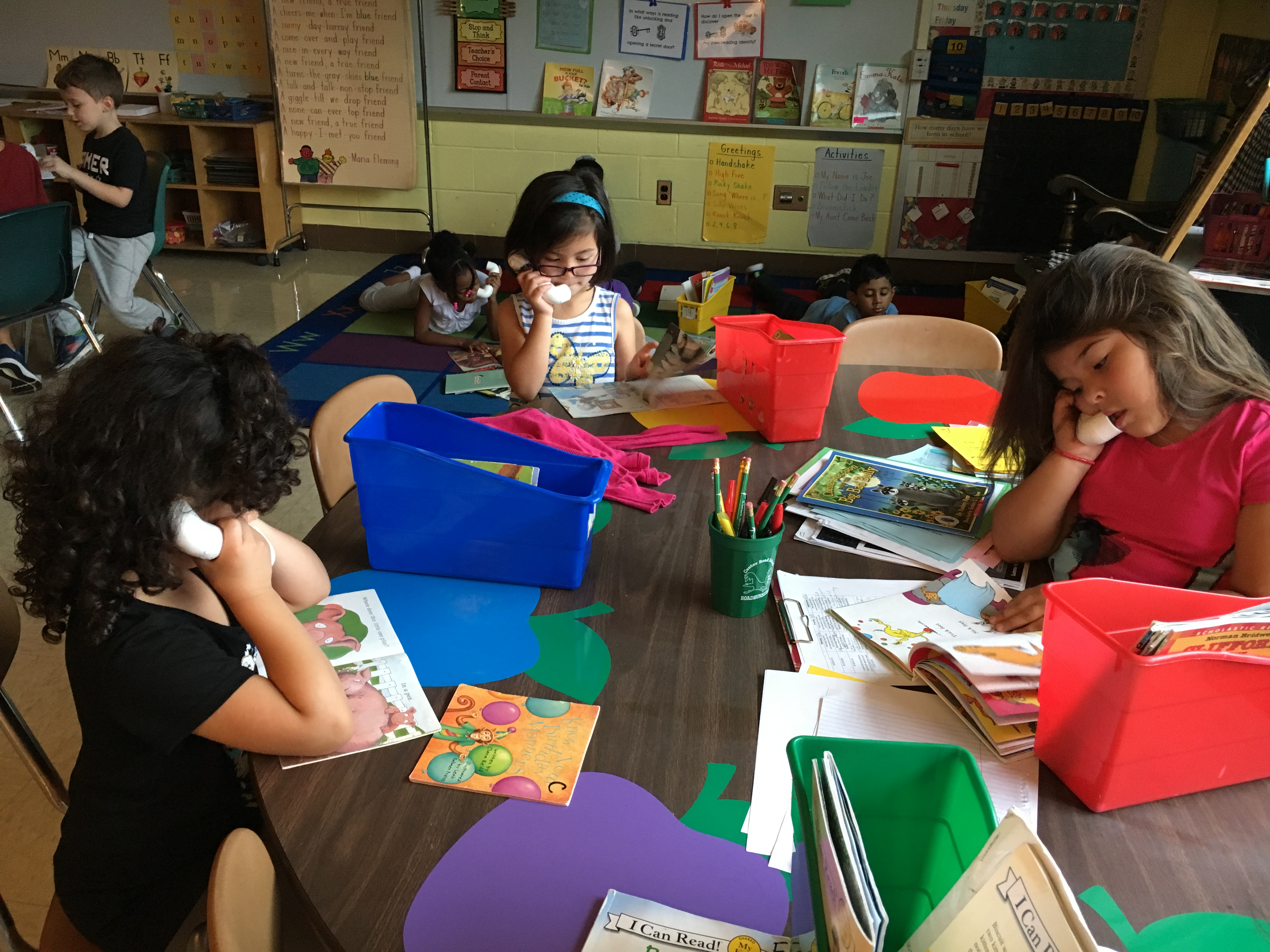 Three children are engaged in activities at a classroom table, with one girl using a toy phone, while others read books and work with colorful materials.