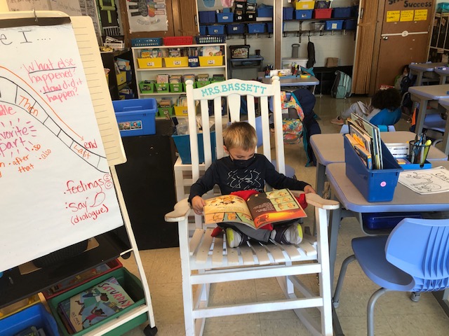 A young child sits in a large white rocking chair, reading a book in a colorful classroom filled with educational materials and furniture.