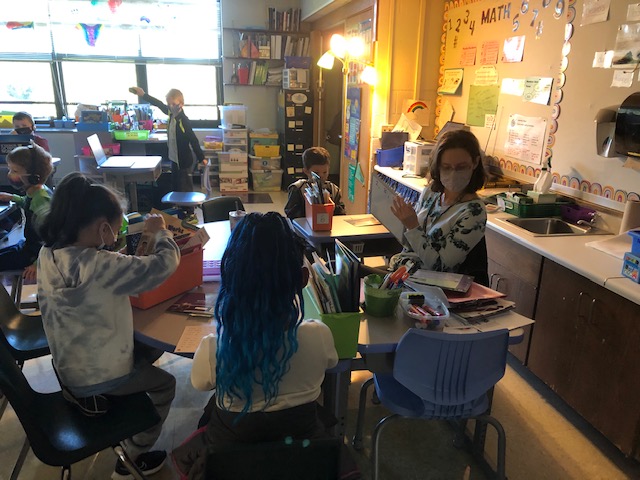 A classroom scene showing several students engaged in activities at their desks, with a teacher interacting with them in a brightly lit environment.