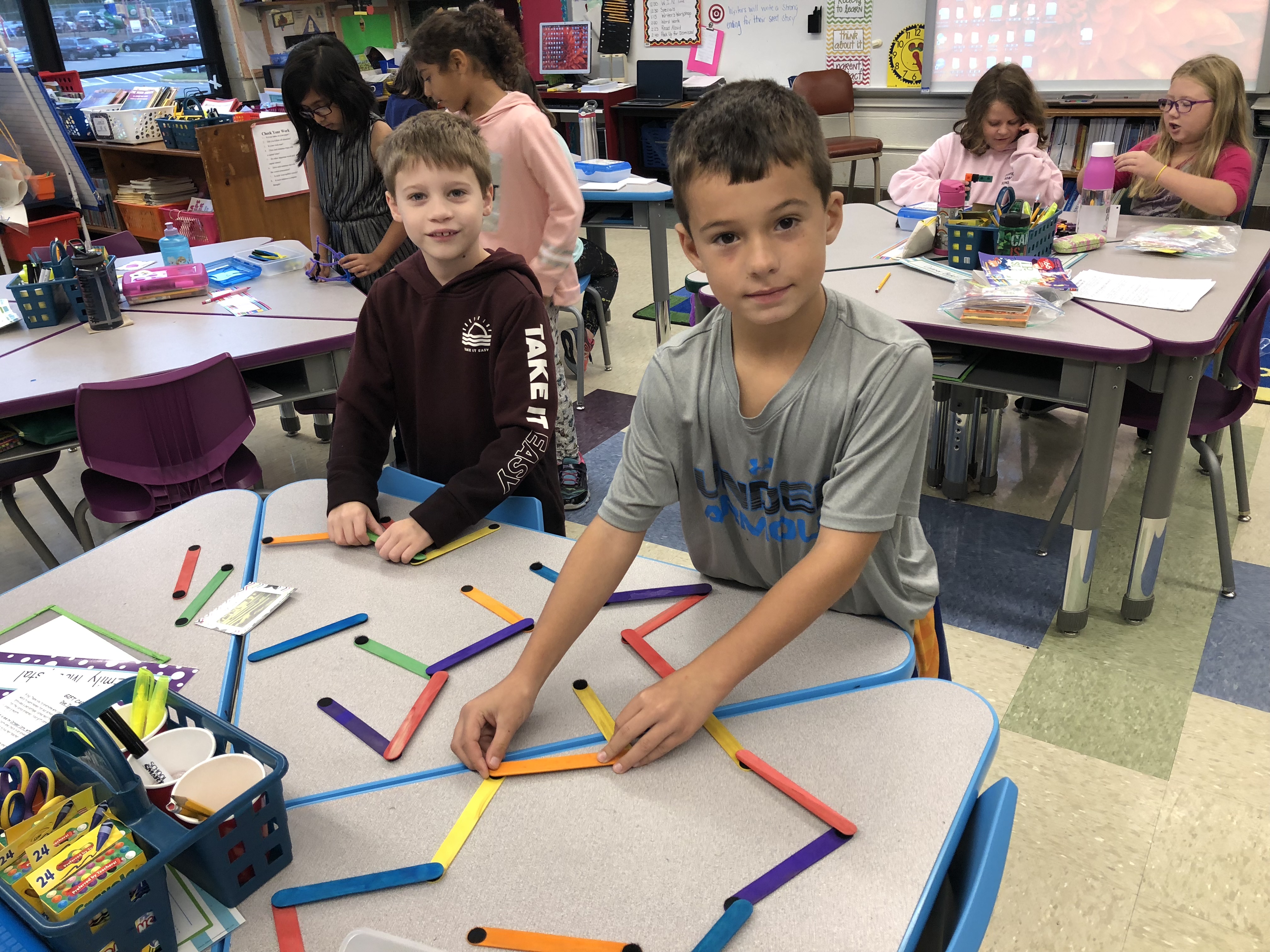 Two children are engaged in a hands-on activity at a classroom table, using colorful sticks to create shapes, while other students work in the background.