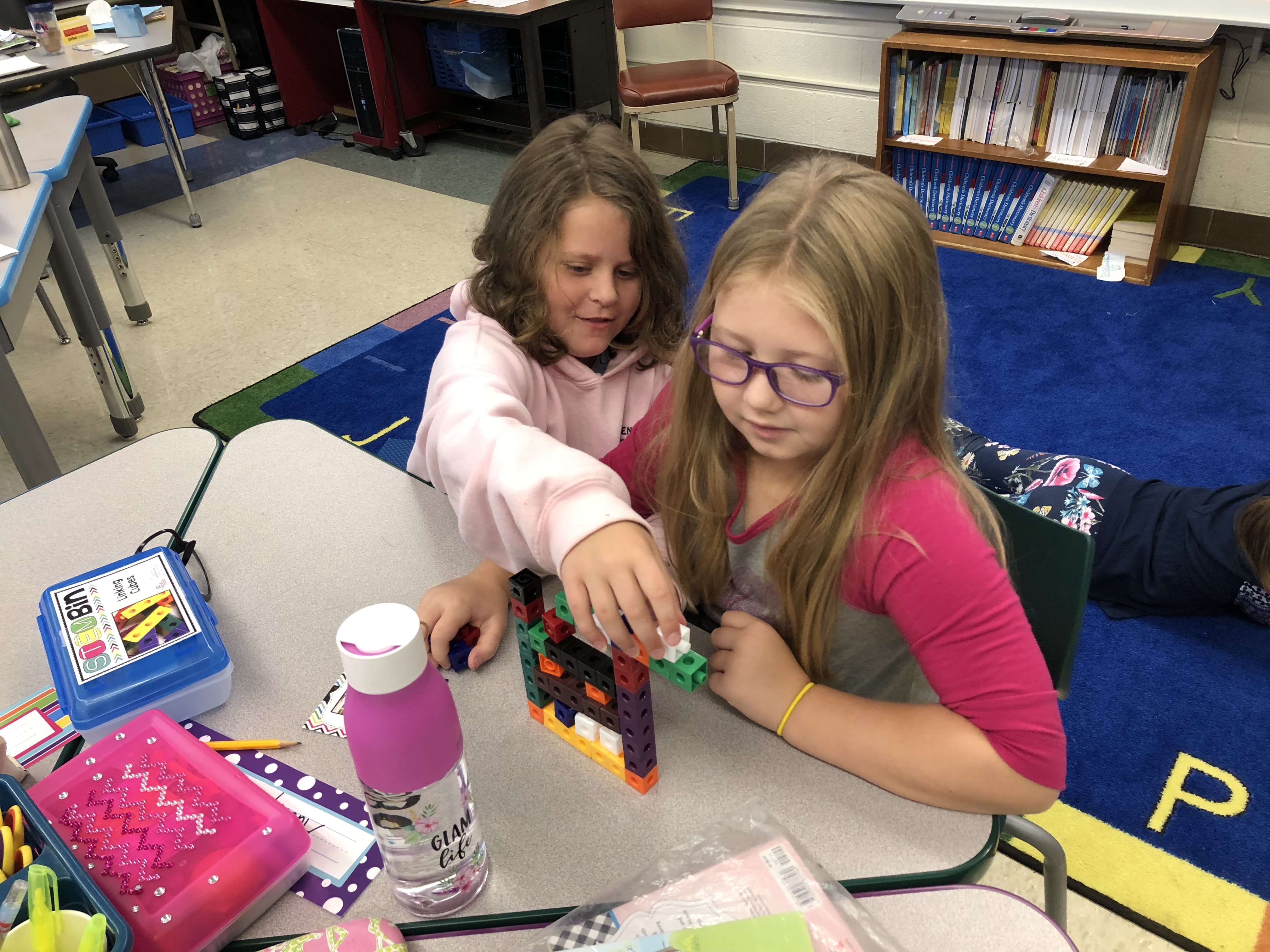Two young girls are collaborating at a classroom table, working together to build a structure with colorful blocks, surrounded by school supplies.