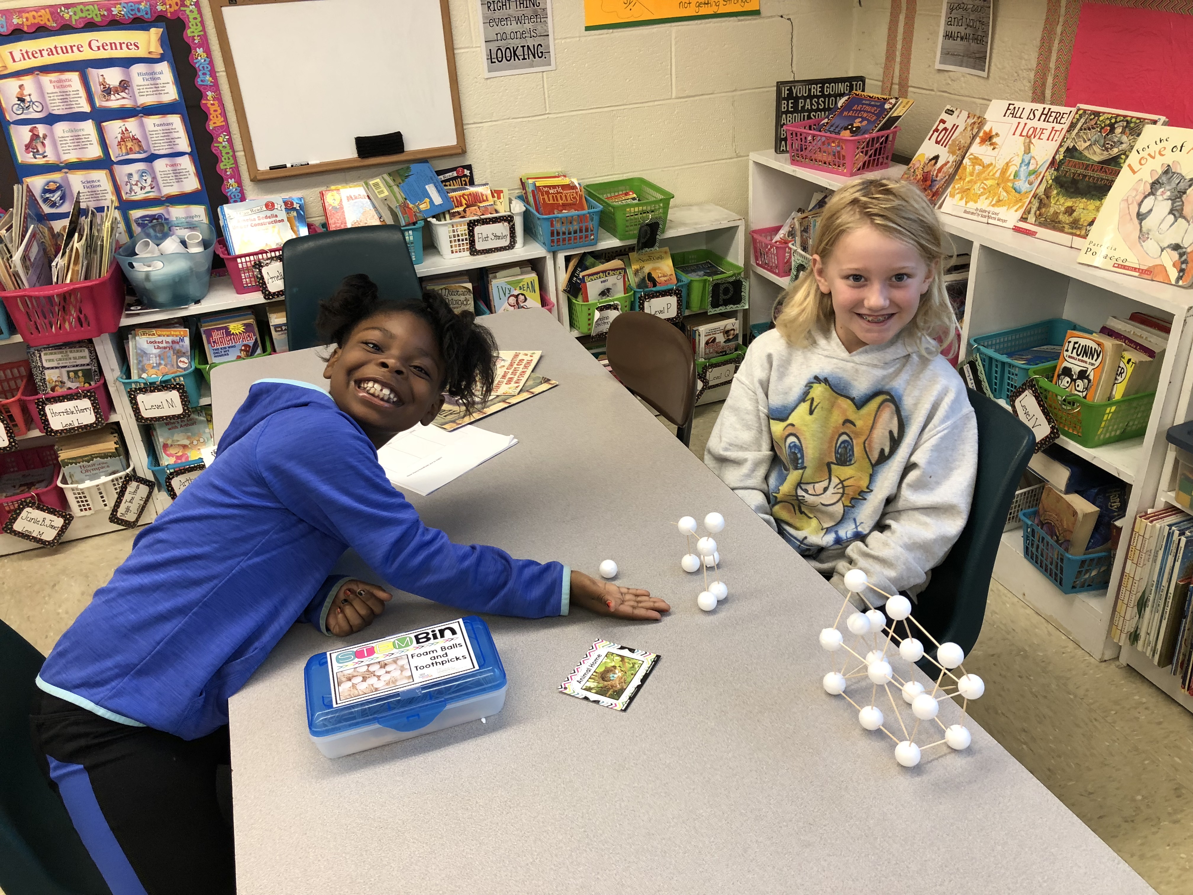 Two children are smiling and engaged in an activity at a classroom table, with educational materials and books visible in the background.