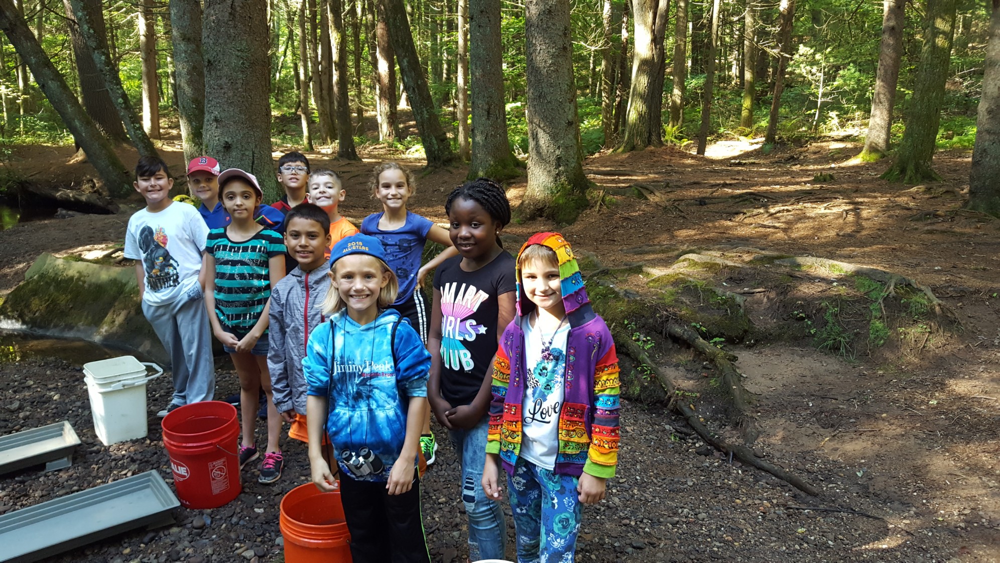 A group of children stands together in a forested area, smiling and wearing colorful clothing, with trees and a path visible in the background.