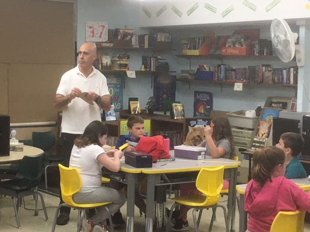 A teacher stands in front of a classroom filled with students engaged in various activities at their desks.