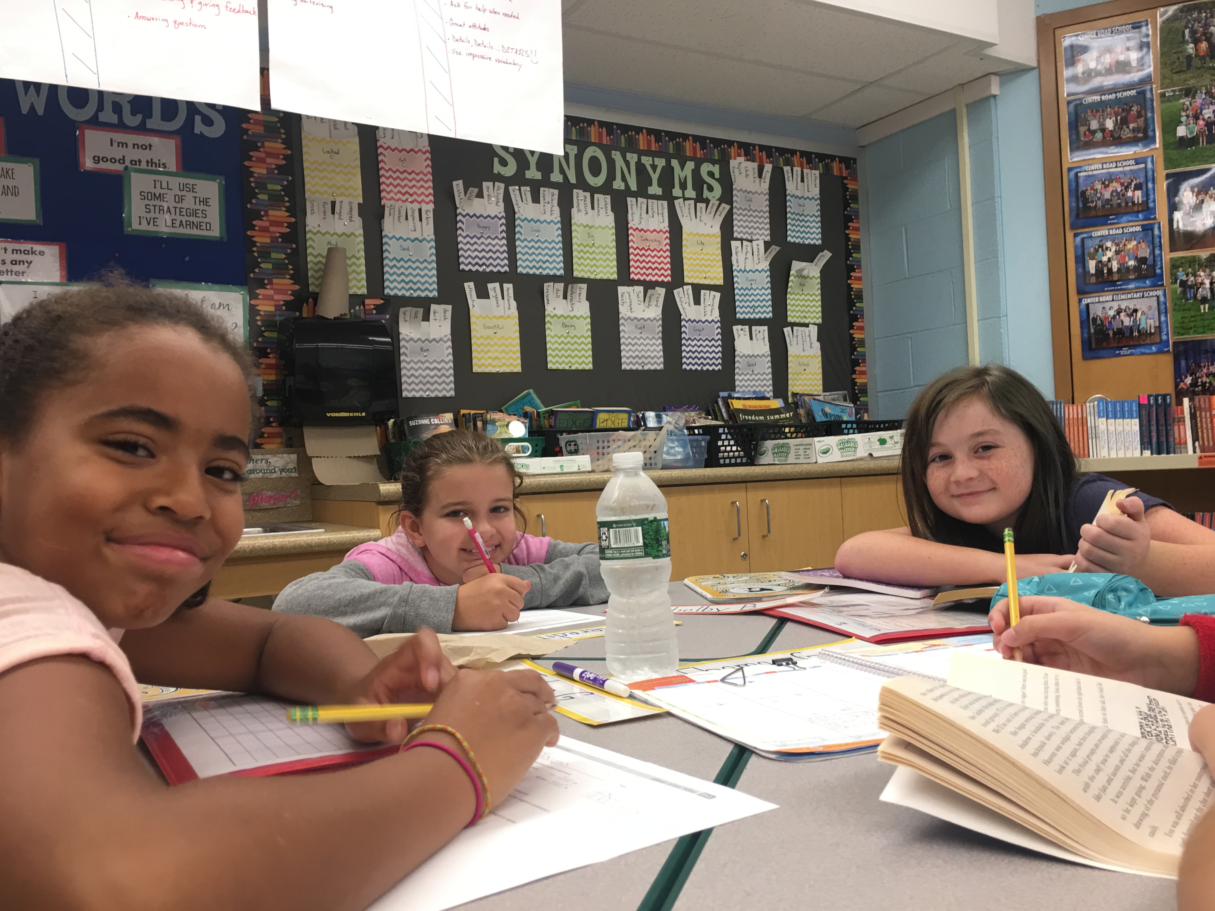 Four children are sitting around a table engaged in a learning activity, with books and writing materials in front of them, and a bulletin board filled with educational materials in the background.